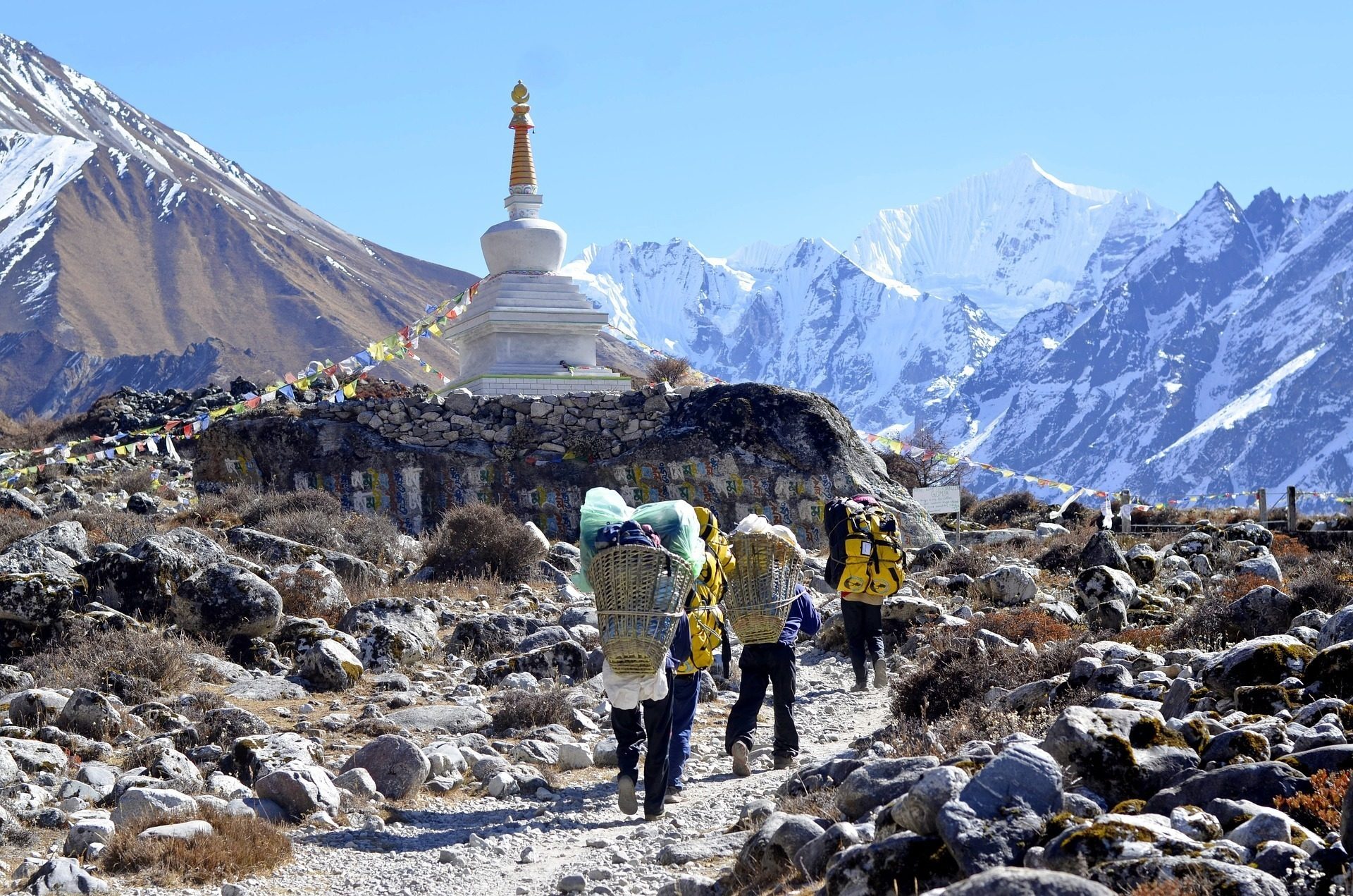 Trekkers and porters on a rocky Himalayan trail passing a Buddhist stupa, with snow peaks behind